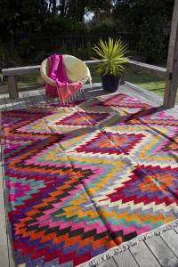 Una alfombra con estampados coloridos, ideal para decorar, dispuesta sobre una terraza de madera con un banco de jardín y una planta en maceta al fondo, y una tela de color rosa brillante cubriendo el banco.