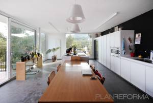 Una moderna cocina con elegantes gabinetes blancos, una larga mesa de comedor de madera y grandes ventanas que ofrecen una vista de un paisaje verde exuberante.