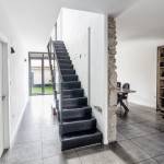 Un pasillo interior moderno de estilo minimalista con una escalera negra a la derecha y grandes baldosas grises; a la izquierda, hay puertas a otras habitaciones y una silla de madera cerca de un mueble alto en