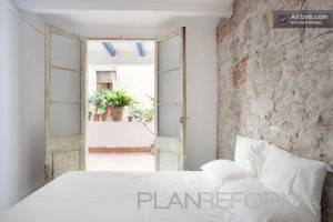 Un dormitorio amplio y luminoso con una puerta abierta que conduce a un pequeño balcón adornado con plantas en macetas. Las paredes parecen tener una textura rústica y la ropa de cama es blanca y fresca. Ideal para aquellos