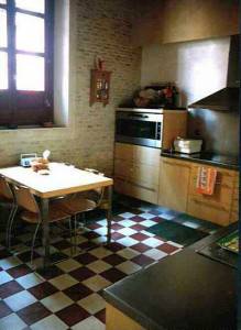 Una modesta cocina con un suelo ajedrezado, ofreciendo una pequeña mesa de comedor, una encimera y electrodomésticos integrados después de su rehabilitación energética.