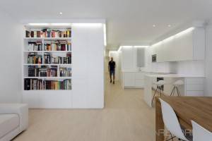 Una habitación moderna y luminosa con una cocina minimalista blanca, reformas en casa como primer paso y una estantería empotrada llena de libros. Hay una silla