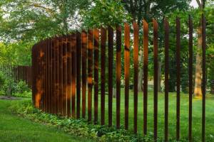 Una valla curva de acero corten de color óxido con listones verticales en un entorno verde y boscoso.