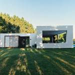 Casa moderna de una sola planta con grandes ventanales de vidrio con vistas a una piscina, con un telón de fondo de árboles y un césped bien cuidado con sombras alargadas que indican la luz del sol temprano en la mañana o al final de la tarde, construida con contenedores ISO