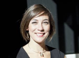 Una mujer sonriente con cabello corto y aretes, de pie en un ambiente tenuemente iluminado con una fuente de luz brillante iluminando un lado de su