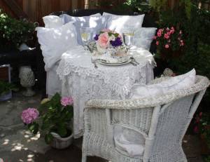 Un pintoresco comedor al aire libre Shabby Chic con una mesa cubierta de encaje para dos personas, vajilla elegante y un jarrón de flores frescas, rodeado de exuberantes plantas de jardín.