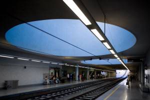 Moderno andén de una estación de metro con pasajeros esperando, con un tragaluz azul ovalado y una gran iluminación lineal.