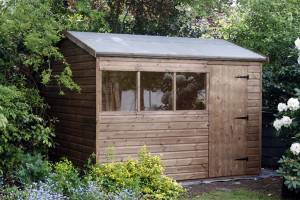Una caseta de jardín de madera para comprar, con una ventana y una puerta, rodeada de vegetación y flores.
