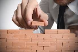 Una persona con camisa coloca un bloque de madera encima de una pared de ladrillos cuidadosamente apilados, simbolizando la construcción o la planificación cuidadosa.