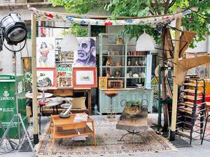 Una exhibición ecléctica al aire libre con una estética vintage, con sillas variadas, una mesa de madera, obras de arte, estantes con varios objetos y una estructura superior decorativa con artículos colgantes en un vibrante barrio decorado.