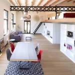 Una espaciosa área de estar moderna con una larga mesa de comedor blanca, sillas rojas, una escalera a la derecha y estantes con diversos objetos decorativos.