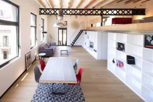 Una espaciosa área de estar moderna con una larga mesa de comedor blanca, sillas rojas, una escalera a la derecha y estantes con diversos objetos decorativos.