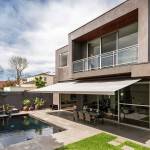 Casa moderna de dos pisos con techo plano, con amplias ventanas de vidrio, balcón y área para sentarse al aire libre, junto a una piscina rectangular y un césped bien cuidado. Ideal para aquellos