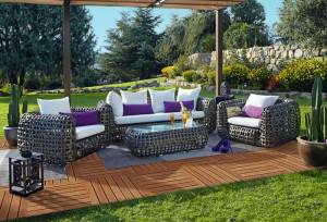 Un patio al aire libre con muebles de mimbre con cojines blancos, almohadas decorativas de color púrpura, una pequeña mesa con tablero de vidrio con una bandeja de bebidas, rodeado por un césped bien cuidado y un jardín bajo un