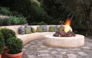 Una zona de estar al aire libre con un brasero circular de piedra que tiene una llama ardiendo en el centro, rodeado por un banco curvo adornado con cojines estampados y de colores sólidos, según los principios del Feng Shui.