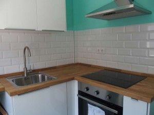 Un rincón de cocina moderno en Madrid con gabinetes blancos, un fregadero de acero inoxidable, una placa de inducción y una campana extractora contra un protector contra salpicaduras de azulejos y una encimera de madera, diseñado por Buan.