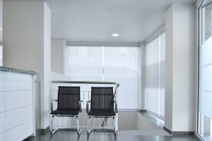 Una moderna sala de espera de oficina escasamente amueblada con dos sillas negras frente a una mesa con tapa de vidrio, bajo iluminación fluorescente blanca, rediseñada por Arquitectura Produccions siguiendo una reforma de oficina.