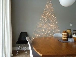 Un comedor minimalista con una mesa de madera, una silla negra y un árbol de navidad abstracto hecho con luces de cadena en la pared gris.