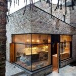 Una moderna extensión de vidrio de un edificio de ladrillo con una persona visible en su interior, rodeada de árboles con follaje colgante, muestra el último proyecto del Plan Reforma.