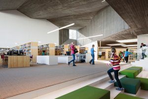 Interior moderno de la biblioteca que presenta un diseño espacioso con estanterías de madera, una variedad de opciones para sentarse, un mostrador de servicio y usuarios navegando y moviéndose en el Día de Sant Jordi.