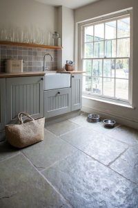 Un diseño de cocina ordenado con suelo de baldosas de piedra, gabinetes grises, una ventana con vistas al exterior, estantes abiertos con cristalería y