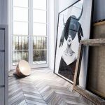 Una obra de arte monocromática que presenta una figura femenina está apoyada contra una pared cerca de una ventana y una puerta, con una lámpara de pie volcada de lado en una habitación con piso de parquet de madera.