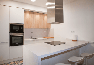Una moderna cocina en Barcelona con mobiliario de madera, encimeras blancas, electrodomésticos integrados y una isla central con asientos.