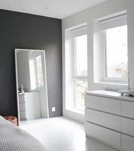Un dormitorio minimalista con paredes grises, molduras blancas, un gran espejo de pie y una cómoda blanca cerca de la ventana en aluminio.