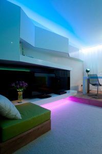 Una moderna sala de estar que presenta iluminación ambiental con tonos azules, una elegante chimenea y muebles contemporáneos que incluyen un otomano.