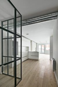 Una cocina moderna con gabinetes y encimeras blancos, con un diseño de planta abierta diseñada por RAS Arquitectura, con conductos expuestos en el techo y pisos de madera clara. Negro con paneles de vidrio