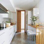Una cocina moderna y estrecha con gabinetes blancos y electrodomésticos de acero inoxidable, que cuenta con una barra desayunadora de madera, luces colgantes, flores decorativas y diseñada para optimizar el costo obra.