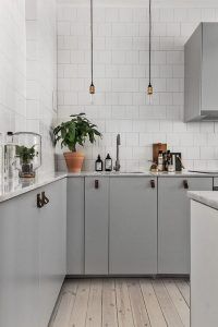 Una cocina moderna con placa para salpicaduras de azulejos blancos, gabinetes grises y pisos de madera clara. Las encimeras se adornan con algunas plantas y accesorios de cocina, elige colores para las luces colgantes.