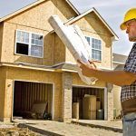 Un trabajador de construcción con casco amarillo está examinando los planos frente a una casa en autopromoción parcialmente terminada.