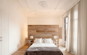 Un dormitorio minimalista con un gran cabecero de madera, paredes blancas, cortinas vaporosas y sencillas mesillas de noche con lámparas pensadas para el descanso.