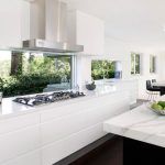 Moderna remodelación de cocina con gabinetes blancos, encimeras de mármol, electrodomésticos de acero inoxidable y una vista de la vegetación a través de la ventana.