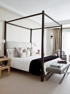 Un dormitorio cuidadosamente organizado con una cama con dosel y ropa de cama blanca y gris, acentuada por almohadas decorativas. Junto a la cama se encuentran dos sillones y una pequeña mesa auxiliar, que llega hasta el suelo.