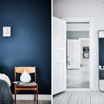 Una habitación moderna con paredes azul oscuro, una mesilla de noche de madera y un lecho crujiente blanco junto a una puerta abierta que conduce.
