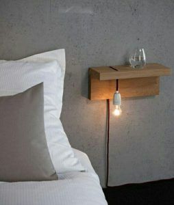 Una estancia minimalista con un estante de madera junto a la cama, con una luz colgante desnuda y un vaso de agua encima, adyacente a una