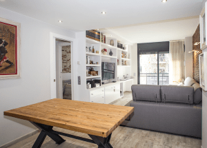 Un salón moderno con una estantería abierta, un televisor montado, una gran mesa de madera en primer plano y un sofá gris frente al televisor. Hay obras de arte en las paredes y luz natural.