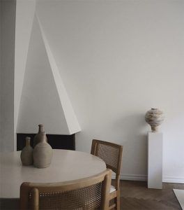 Una habitación minimalista con una mesa redonda, dos jarrones de cerámica sobre la mesa, un jarrón más grande sobre un pedestal blanco típico del estilo madrileño, una sola silla y un patrón de luces geométrico en la pared.