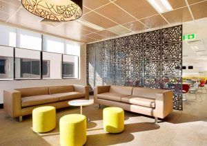 Una moderna sala de estar de oficina con sofás de colores neutros, otomanas amarillas y un separador de ambientes negro ornamentado, con abundante luz natural.