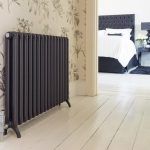 Una instalación de radiadores tradicionales frente a una pared empapelada con motivos florales, con vista a un dormitorio que muestra una cama con cabecero oscuro de fondo.