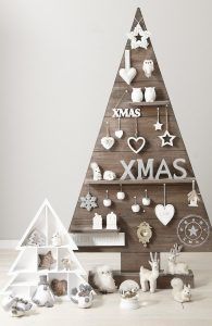 Un estante triangular de madera hecho a mano dispuesto en forma de árbol de Navidad, decorado con adornos festivos y acompañado de adornos más pequeños de temática navideña sobre un fondo neutro.