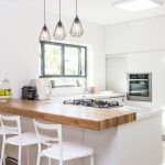 Interior de cocina moderno con gabinetes blancos, electrodomésticos de acero inoxidable, una encimera de madera con taburetes blancos e iluminación colgante para lograr una cocina funcional.