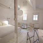 Una habitación minimalista blanca que presenta una cama de dos plazas, dos pequeñas ventanas con contraventanas, lámparas de pie, un radiador, un