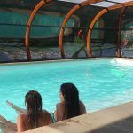Dos individuos relajándose en una piscina interior con cubiertas para piscinas transparentes, de arco y una vista del verdor circundante.
