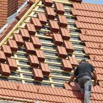Un trabajador está instalando tejas rojas en unos tejados residenciales empinados con algunas secciones ya terminadas.