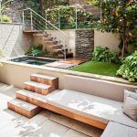 Una acogedora terraza urbana que cuenta con una pequeña piscina, madera para la plataforma con escalones, césped artificial, exuberantes plantas en macetas.