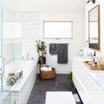 Un baño moderno con azulejos blancos del metro, una mampara de ducha de vidrio, un espejo con marco de madera y azulejos del suelo del baño gris oscuro. Los accesorios incluyen una planta en maceta y toallas en una cesta.