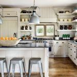 Una moderna cocina interior que destaca por su blanca en la carpintería, estantes abiertos, una isla central con taburetes de bar y lámparas col.